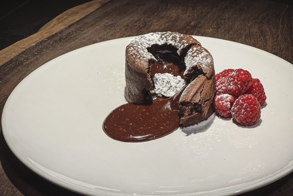 Σοκολατένιο γλυκό Fondant κομμένο στη μέση με ρευστή σοκολάτα να τρέχει, διακοσμημένο με άχνη ζάχαρη και σμέουρα