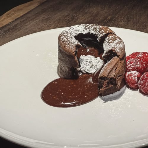 Σοκολατένιο γλυκό Fondant κομμένο στη μέση με ρευστή σοκολάτα να τρέχει, διακοσμημένο με άχνη ζάχαρη και σμέουρα